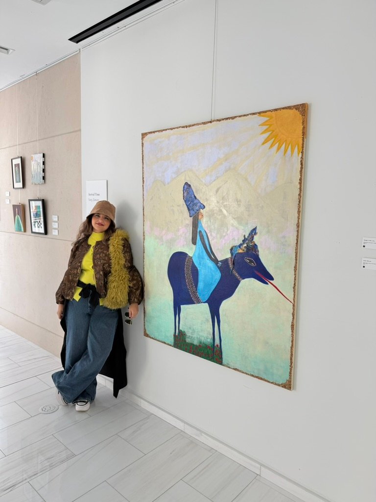 Woman standing beside a painting of a figure in blue riding a blue animal under a yellow sun at the Unpromised Flowers exhibition