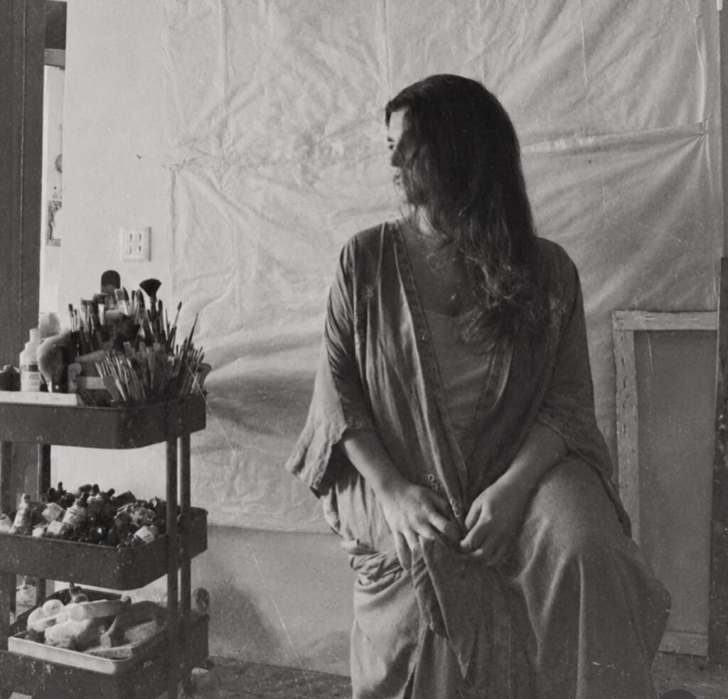 Nazende Afakoğlu sitting in her art studio, turned away from the camera, beside a trolley full of paintbrushes and paint tubes, in black and white